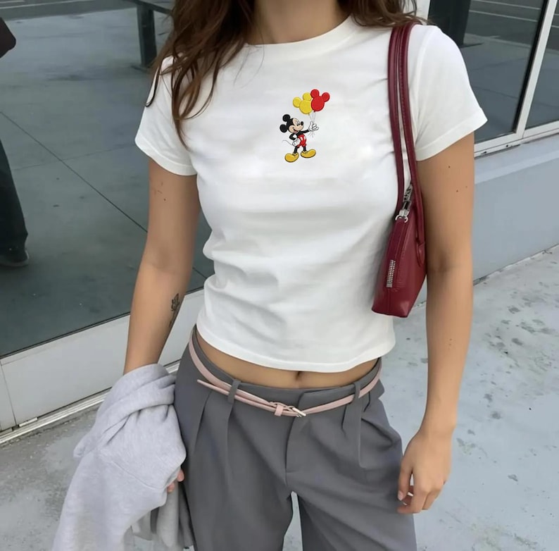 Op de afbeelding: Een wit T-shirt met korte mouwen en een kleine afbeelding van Mickey Mouse die drie gele en rode ballonnen vasthoudt. Het shirt wordt gecombineerd met een grijze broek met een dunne roze riem en een donkerrode schoudertas met smalle band.