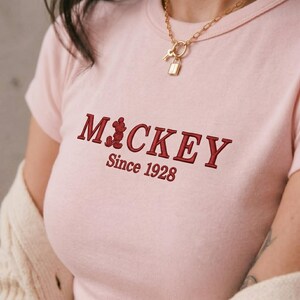 May include: A light pink short-sleeved t-shirt featuring dark red embroidered text that reads MICKEY with a small silhouette of Mickey Mouse replacing the letter I, followed by the text Since 1928 underneath in a matching dark red font.