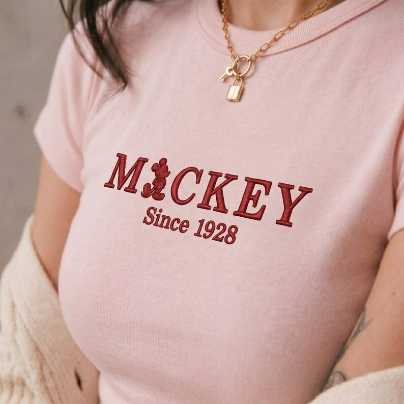 May include: A light pink short-sleeved t-shirt featuring dark red embroidered text that reads MICKEY with a small silhouette of Mickey Mouse replacing the letter I, followed by the text Since 1928 underneath in a matching dark red font.