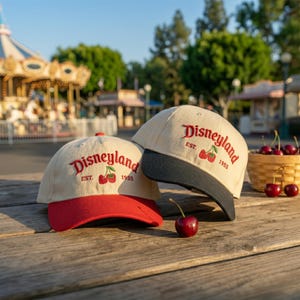 Op de afbeelding: Twee baseballpetten met crèmekleurige kronen en geborduurde rode tekst Disneyland EST. 1955 boven een paar rode kersen. Eén pet heeft een rode klep, de andere een zwarte klep. Er ligt een losse kers naast de petten op de houten ondergrond.