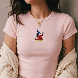 May include: A light pink short-sleeved t-shirt featuring a small embroidered graphic of a cartoon mouse wearing a blue wizard hat and a red robe, making a peace sign gesture. The shirt has a crew neckline and is paired with a gold chain necklace.