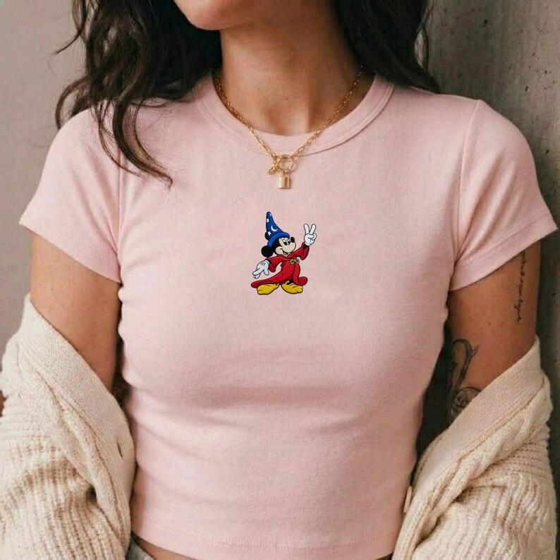 May include: A light pink short-sleeved t-shirt featuring a small embroidered graphic of a cartoon mouse wearing a blue wizard hat and a red robe, making a peace sign gesture. The shirt has a crew neckline and is paired with a gold chain necklace.