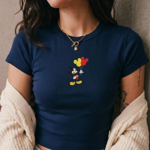 Op de afbeelding: Een marineblauw t-shirt met korte mouwen en een kleine geborduurde afbeelding van Mickey Mouse die een gele en een rode ballon vasthoudt. Het shirt heeft een ronde hals, een geribbelde textuur en wordt gedragen met een goudkleurige ketting.