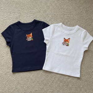 May include: Two short-sleeved crop tops, one navy blue and one white. Each shirt features an embroidered image of a fox and a rabbit. The shirts are made of a soft material and are suitable for casual wear.