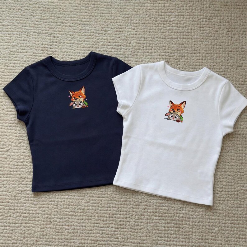 May include: Two short-sleeved crop tops, one navy blue and one white. Each shirt features an embroidered image of a fox and a rabbit. The shirts are made of a soft material and are suitable for casual wear.