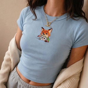 May include: Light blue crop top featuring a cartoon image of a fox and a rabbit. The fox is orange and the rabbit is gray. The top has short sleeves and a round neckline. The image is centered on the front of the shirt.