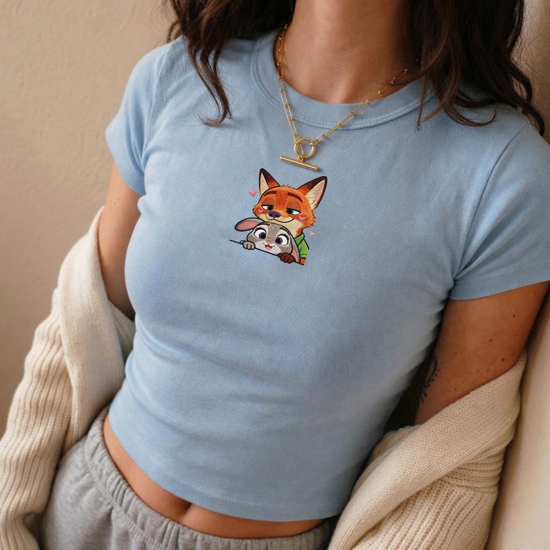 May include: Light blue crop top featuring a cartoon image of a fox and a rabbit. The fox is orange and the rabbit is gray. The top has short sleeves and a round neckline. The image is centered on the front of the shirt.