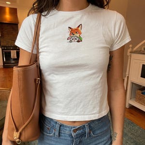 May include: White crew neck t-shirt featuring a colorful cartoon image of a fox and a rabbit. The shirt is a short-sleeved style and is paired with blue jeans. A brown leather tote bag is visible on the left side of the image.