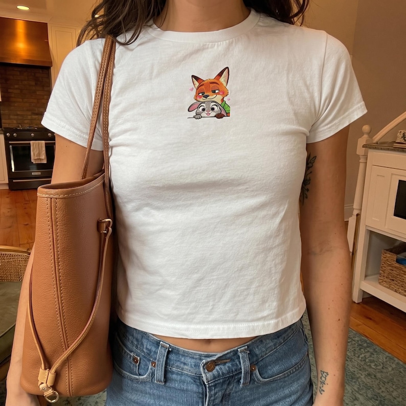 May include: White crew neck t-shirt featuring a colorful cartoon image of a fox and a rabbit. The shirt is a short-sleeved style and is paired with blue jeans. A brown leather tote bag is visible on the left side of the image.