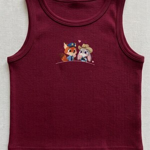 May include: A burgundy ribbed tank top with a crew neck. The tank top features an embroidered design of a fox and a bunny, both wearing hats. The fox is wearing a police hat, and the bunny is wearing a cowboy hat. Two small pink hearts are above the characters.