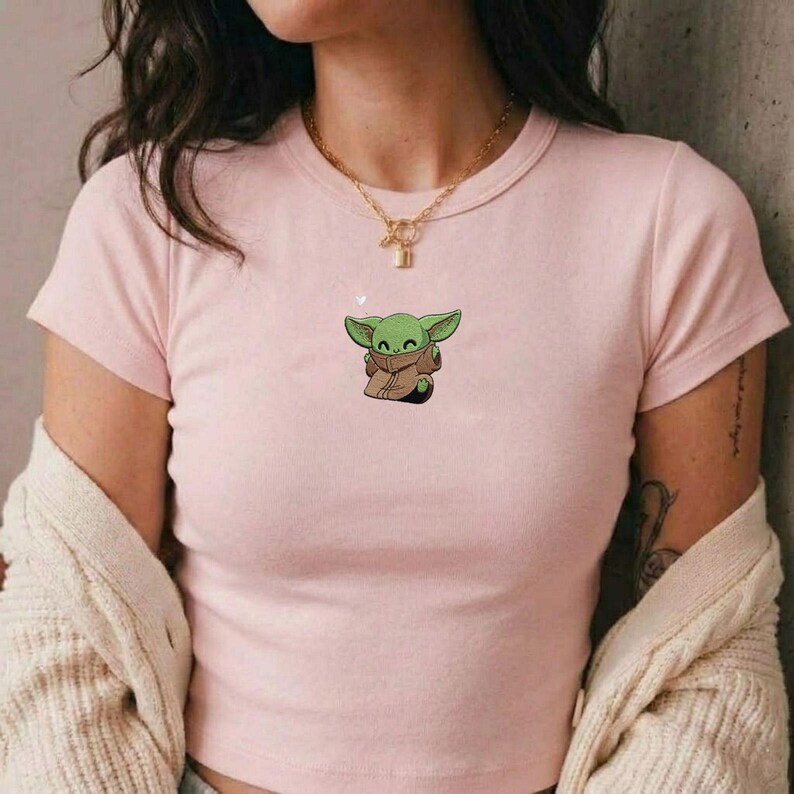 May include: A light pink short-sleeved t-shirt featuring a small, centered graphic of a green, large-eared creature wearing a brown robe. The shirt has a crew neckline and a fitted silhouette, paired with a gold-toned chain necklace.