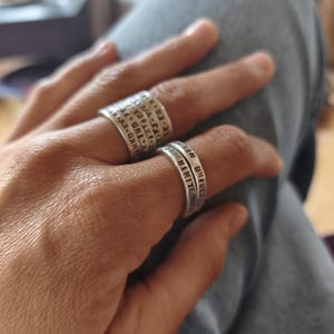 Peut inclure: Deux bagues en argent sont présentées sur une main. L'une est large avec des symboles gravés, l'autre est une bande plus fine avec le mot "UNIVERSE" gravé. Les bagues sont portées sur une main, avec un arrière-plan flou.