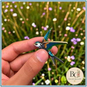Peut inclure: Une broche en forme d'oiseau avec des ailes en émail bleu et vert vif aux bords en métal doré. L'oiseau présente une poitrine blanche, une tête sombre et une longue queue pointue. La pièce est ornée de détails métalliques et d'émail coloré.