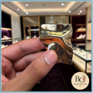 May include: A gold-toned metal brooch with a polished, reflective surface and an irregular, wavy, organic shape. The piece features smooth, flowing curves and a high-shine finish that catches the light, creating deep shadows and bright highlights.