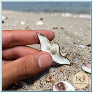Könnte beinhalten: Eine goldgeränderte Brosche in Form einer geschwungenen, länglichen Muschel mit glatter weißer Emaille-Oberfläche. Zwei kleine, runde, weiße Perlen sind in der Mitte der muschelartigen Form eingesetzt, die einen polierten Metallrand aufweist.