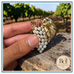 Op de afbeelding: Een goudkleurige metalen broche in de vorm van een tros druiven, voorzien van meerdere ronde witte kunstparels in een driehoekige opstelling, geaccentueerd door een gestructureerd gouden blad en een gekruld rankdetail bovenaan.