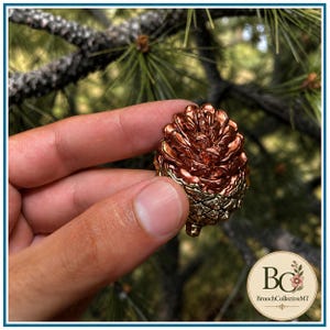 Puede incluir: Un broche metálico con forma de piña que presenta un diseño bicolor con una sección superior de color cobre pulido y una base texturizada en tono oro antiguo. La pieza muestra escamas superpuestas que imitan la estructura natural.