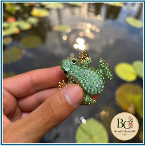 Könnte beinhalten: Eine froschförmige Brosche, die mit kleinen hellgrünen Kristallen besetzt ist. Der Frosch hat zwei dunkle, runde Augen sowie Beine und Füße aus goldfarbenem Metall. Der Körper ist dicht mit funkelnden grünen Strasssteinen bedeckt.