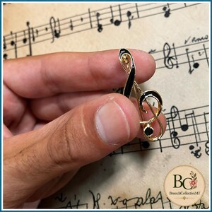 Könnte beinhalten: Eine goldfarbene Metallbrosche in Form eines Violinschlüssels mit glatter schwarzer Emaille-Einlage entlang der geschwungenen Linien und einem kleinen, runden schwarzen Steinakzent an der Basis. Die Brosche hat ein poliertes Finish.