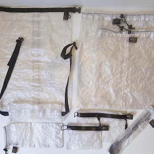 May include: A collection of translucent white, crinkled material storage bags and pouches. The set includes a large roll-top dry bag with black straps and buckles, two drawstring bags, two zippered pouches, and a long, narrow sleeve with a drawstring.