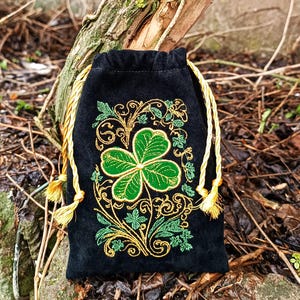 May include: Black velvet drawstring bag with a green shamrock and gold embroidery. The shamrock is the focal point, surrounded by intricate gold and green floral designs. The bag has gold drawstrings with tassels.