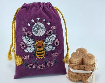 May include: A purple velvet drawstring pouch featuring an embroidered design of a bee, a moon, stars, and pink flowers, accompanied by three cylindrical beeswax candles wrapped in burlap and tied with a white ribbon.