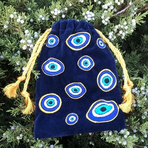 Op de afbeelding: Een donkerblauw fluwelen buideltje met trekkoord, voorzien van meerdere geborduurde boze oog-motieven in tinten blauw, wit en zwart. Het tasje wordt dichtgetrokken met twee dikke, goudgele gedraaide koorden die eindigen in bijpassende kwastjes.