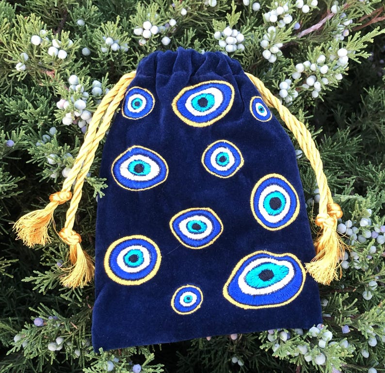 Op de afbeelding: Een donkerblauw fluwelen buideltje met trekkoord, voorzien van meerdere geborduurde boze oog-motieven in tinten blauw, wit en zwart. Het tasje wordt dichtgetrokken met twee dikke, goudgele gedraaide koorden die eindigen in bijpassende kwastjes.