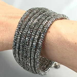 Puede incluir: Una pulsera de múltiples hilos que presenta varias filas de cuentas de color gris oscuro, facetadas y similares a cristales, que crean una apariencia texturizada y brillante. La pulsera envuelve la muñeca, mostrando una disposición densa y uniforme de pequeñas piedras reflectantes.