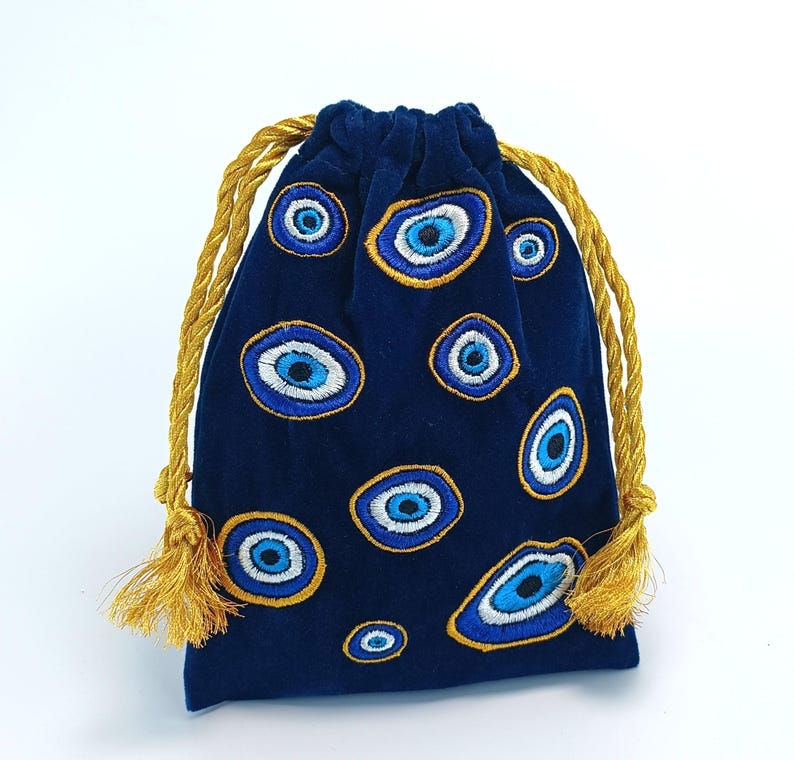 Op de afbeelding: Een donkerblauw fluwelen buideltje met trekkoord, voorzien van een geborduurd patroon van meerdere boze oog-motieven in blauwe, witte en gouden tinten. De tas is bovenaan dichtgetrokken met twee dikke, gedraaide gouden koorden met bijpassende kwastjes.