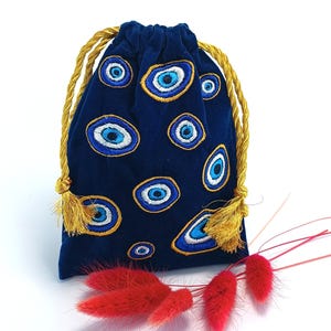 Op de afbeelding: Een donkerblauw fluwelen trekkoordzakje met een geborduurd patroon van meerdere boze oog-motieven in blauwe, witte en gouden tinten. De tas wordt gesloten met twee gedraaide goudkleurige koorden die eindigen in kleine decoratieve kwastjes.