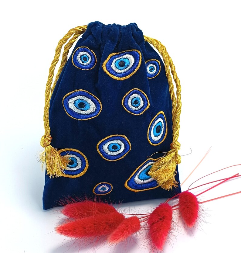 Op de afbeelding: Een donkerblauw fluwelen trekkoordzakje met een geborduurd patroon van meerdere boze oog-motieven in blauwe, witte en gouden tinten. De tas wordt gesloten met twee gedraaide goudkleurige koorden die eindigen in kleine decoratieve kwastjes.