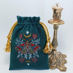 May include: A dark teal drawstring pouch featuring an embroidered moth with red and blue wings, surrounded by white foliage and red berries. Above and below the moth are gold crescent moons and white stars. A metallic candle holder stands nearby.