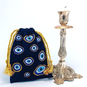 Op de afbeelding: Een donkerblauw fluwelen buideltje met trekkoord, voorzien van geborduurde boze oog-motieven in blauw, wit en goud, naast een hoge, sierlijke kandelaar van zilverkleurig metaal met gedetailleerde bloem- en bladreli&euml;fs.