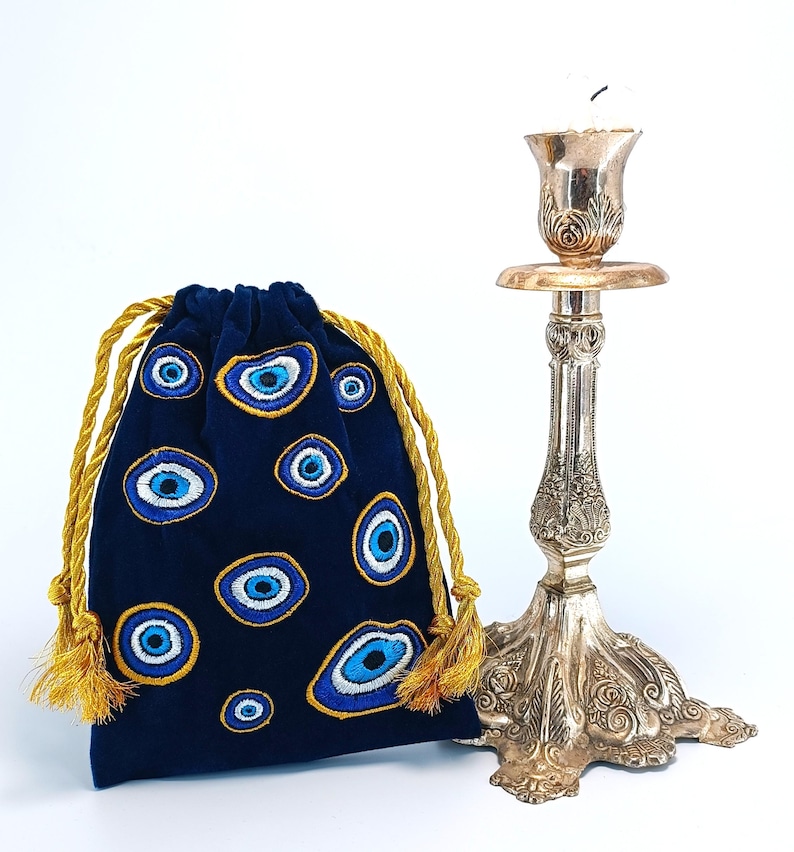 Op de afbeelding: Een donkerblauw fluwelen buideltje met trekkoord, voorzien van geborduurde boze oog-motieven in blauw, wit en goud, naast een hoge, sierlijke kandelaar van zilverkleurig metaal met gedetailleerde bloem- en bladreli&euml;fs.