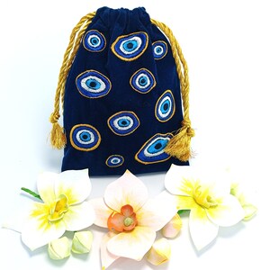 Op de afbeelding: Een donkerblauw fluwelen buideltje met trekkoord, voorzien van een geborduurd patroon van meerdere boze oog-symbolen in blauwe, witte en gouden tinten. De tas wordt gesloten met twee gouden gedraaide koorden met kleine kwastjes aan de uiteinden.