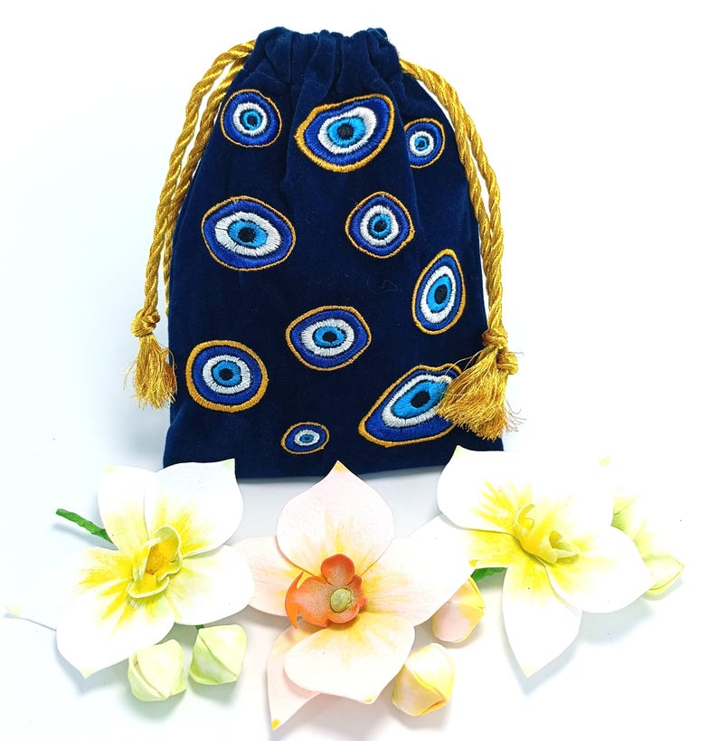 Op de afbeelding: Een donkerblauw fluwelen buideltje met trekkoord, voorzien van een geborduurd patroon van meerdere boze oog-symbolen in blauwe, witte en gouden tinten. De tas wordt gesloten met twee gouden gedraaide koorden met kleine kwastjes aan de uiteinden.
