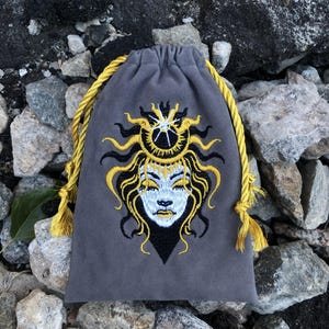Puede incluir: Una bolsa gris con cordón que presenta un diseño bordado de un rostro con los ojos cerrados, enmarcado por cabello fluido y una corona similar a un sol en hilos amarillo, negro y blanco. La bolsa se asegura con cordones trenzados amarillo brillante.