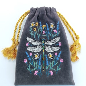 May include: A charcoal gray drawstring pouch featuring an embroidered dragonfly centered among colorful wildflowers. The bag is crafted from a soft, suede-like material and includes two golden yellow twisted cord drawstrings with tasseled ends.
