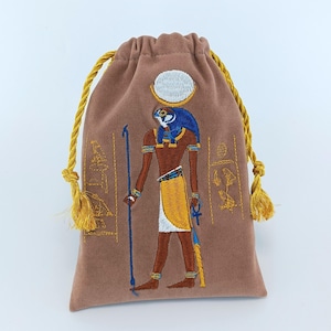 以下が含まれることがあります： ハヤブサの頭、太陽円盤、杖を持つエジプトの神が刺繍された、ライトブラウンの巾着袋。黄色、青、白の糸で細部まで表現されており、両サイドには縦に並んだヒエログリフの刺繍が施されています。