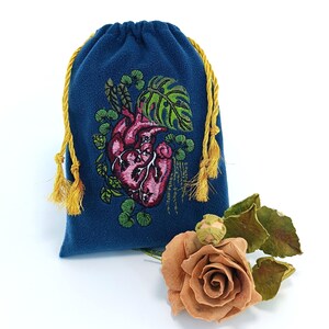 May include: A dark blue drawstring pouch featuring an embroidered anatomical heart surrounded by green monstera and trailing leaves. The bag has two yellow braided cords with tassels at the ends. A dried peach-colored rose sits nearby.