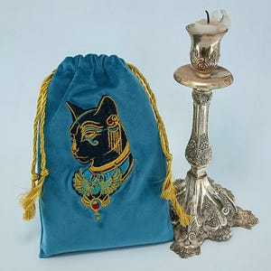 May include: A teal velvet drawstring pouch featuring an embroidered black cat head with gold and blue Egyptian-style jewelry, positioned next to a tall, ornate silver-toned metal candlestick holding a small, partially melted white candle.