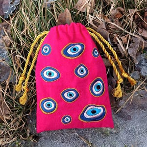 Könnte beinhalten: Ein pinkfarbener Kordelzugbeutel mit Stickereien von blauen und weißen bösen Augen. Der Beutel hat einen goldenen Kordelverschluss mit Quasten. Der Beutel besteht aus einem weichen, plüschigen Material.