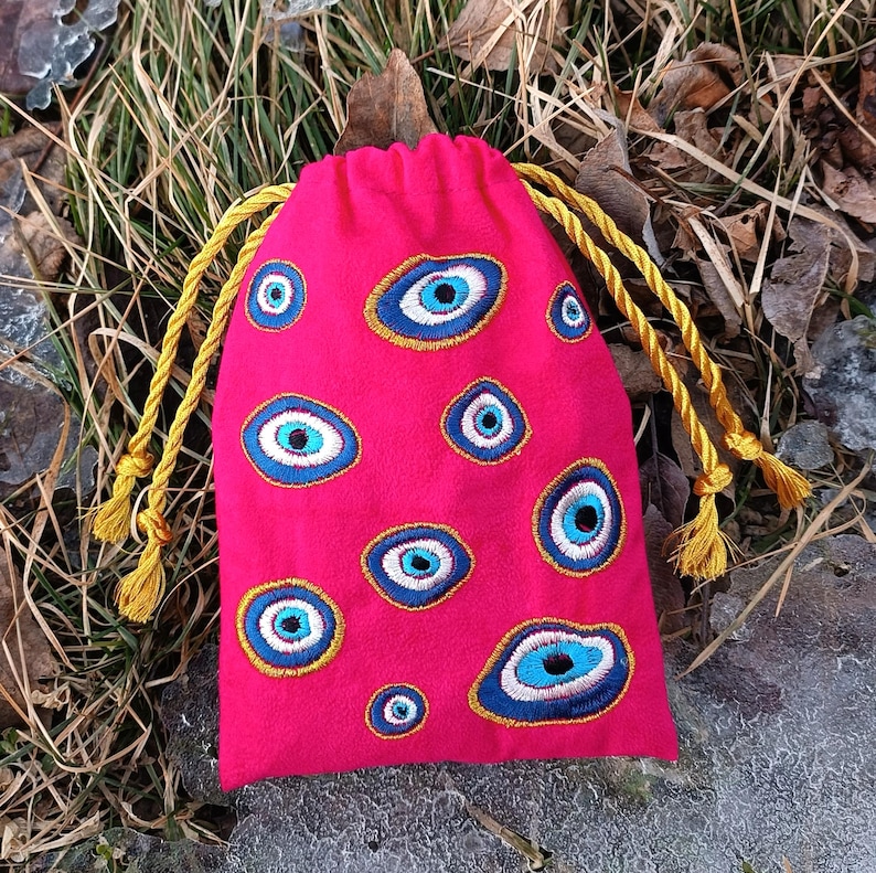 Op de afbeelding: Een felroze buidel met trekkoord, voorzien van meerdere geborduurde boze oog-motieven in blauwe, witte en zwarte tinten, met goudkleurige draadcontouren en bijpassende geelgouden gedraaide koorden bij de sluiting aan de bovenzijde.