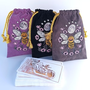 Könnte beinhalten: Drei Samtbeutel mit Kordelzug in Lila, Schwarz und Grau, jeweils mit einer Stickerei aus Biene, Mond und Blumen sowie goldenen Kordeln. Vor den Beuteln liegt ein Tarotkartendeck, wobei die Karte Königin der Kelche oben auf dem Stapel zu sehen ist.