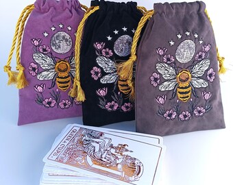 May include: Three velvet drawstring pouches in purple, black, and grey, each featuring an embroidered bee, moon, and floral design with gold cords. A deck of tarot cards is placed in front, with the Queen of Cups card visible on top of the stack.