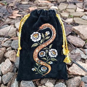 Puede incluir: Una bolsa de terciopelo negro con cordón ajustable que presenta un diseño bordado de una serpiente de color cobre que se enrosca entre flores blancas y hojas verdes. La bolsa se cierra con dos cordones trenzados amarillo dorado.