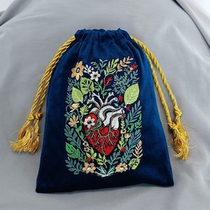 May include: A deep blue velvet drawstring pouch featuring an intricate embroidered design of an anatomical heart surrounded by various colorful flowers and green leaves. The bag is cinched with two thick, twisted gold-colored cord drawstrings.