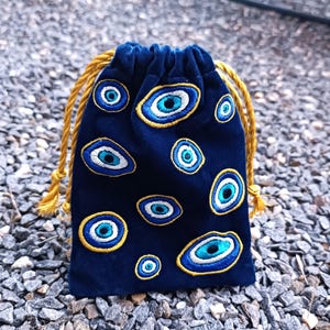 Op de afbeelding: Een donkerblauw fluwelen buideltje met trekkoord, voorzien van meerdere geborduurde boze oog-motieven in blauwe, witte en gouden tinten. De tas wordt aan de bovenkant dichtgetrokken met twee dikke, gedraaide goudgele koorden.
