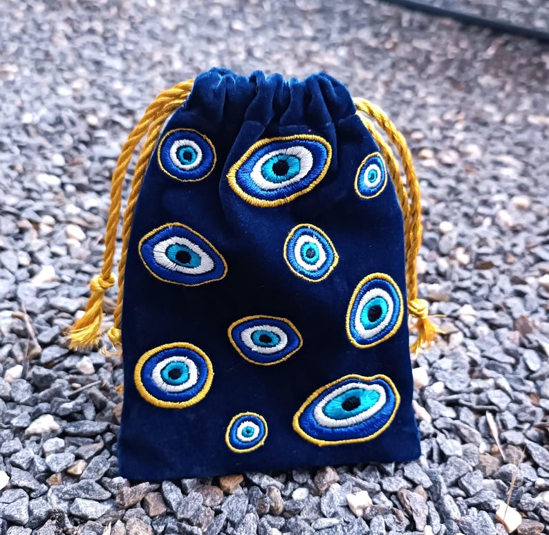 Op de afbeelding: Een donkerblauw fluwelen buideltje met trekkoord, voorzien van meerdere geborduurde boze oog-motieven in blauwe, witte en gouden tinten. De tas wordt aan de bovenkant dichtgetrokken met twee dikke, gedraaide goudgele koorden.